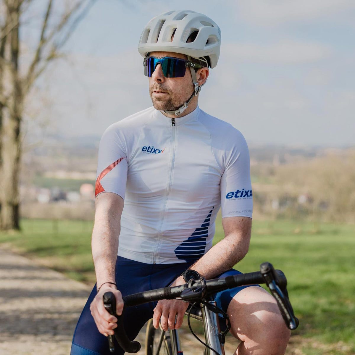 Cyclist wearing a white Etixx cycling jersey with blue and red details, sitting on a road bike in a rural landscape.